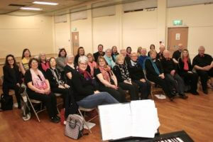 Photo of a group of singers sitting down in rows and smiling at the camera, with a keyboard in the foreground