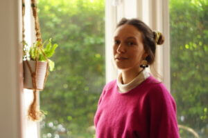 Photo of Hannah in front a window with greenery outside and a hanging basket with plant to the left of the image